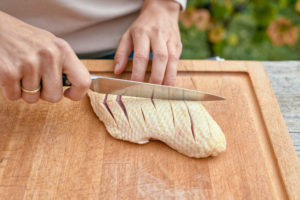 Frauenhände mit Kochmesser ritzen die Haut einer Entenbrustein, Holzbrett Frauenhände mit Kochmesser ritzen die Haut einer Entenbrustein, Holzbrett